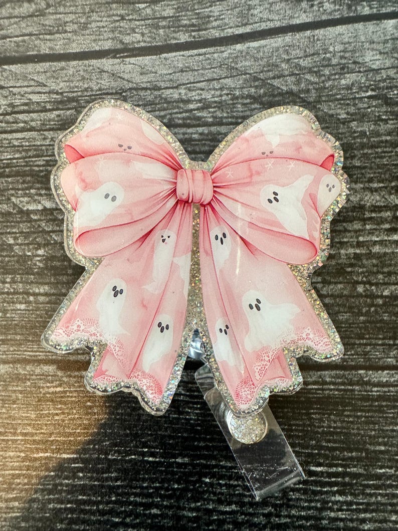 May include: A pink glitter badge holder with a large pink bow and white ghost silhouettes. The bow has a white lace trim.