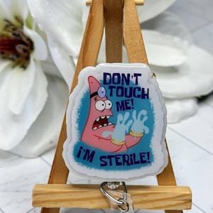 May include: A badge reel featuring a cartoon character with pink skin, wearing a head mirror and white gloves, set against a blue background. The text reads DON'T TOUCH ME! I'M STERILE! The design is encased in a clear, glossy, die-cut acrylic shape.