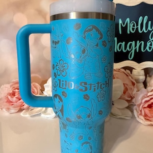 May include: A bright blue insulated tumbler with a handle and a white straw. The exterior features a repeating laser-etched pattern of Lilo and Stitch characters surrounded by hibiscus flowers, with the text Lilo & Stitch printed in the center.