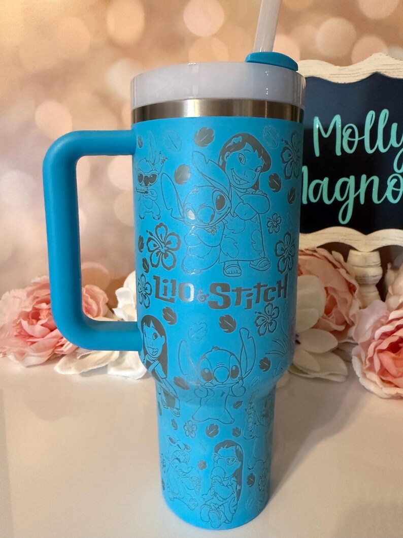 May include: A bright blue insulated tumbler with a handle and a white straw. The exterior features a repeating laser-etched pattern of Lilo and Stitch characters surrounded by hibiscus flowers, with the text Lilo & Stitch printed in the center.