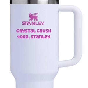 Puede incluir: Un vaso t&eacute;rmico blanco con asa, tapa transparente y pajita transparente. El frente presenta un logotipo rosa de un oso con alas sobre el texto STANLEY, CRYSTAL CRUSH, 40OZ. STANLEY. El vaso tiene una base c&oacute;nica y un acabado mate suave.