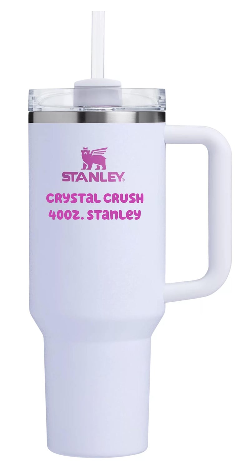 Puede incluir: Un vaso t&eacute;rmico blanco con asa, tapa transparente y pajita transparente. El frente presenta un logotipo rosa de un oso con alas sobre el texto STANLEY, CRYSTAL CRUSH, 40OZ. STANLEY. El vaso tiene una base c&oacute;nica y un acabado mate suave.