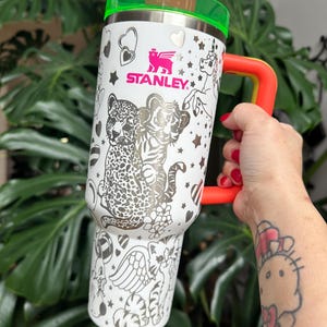 Puede incluir: Un vaso Stanley blanco con una tapa verde ne&oacute;n vibrante, un asa naranja brillante y una pajita a juego de color verde ne&oacute;n. El exterior est&aacute; decorado con ilustraciones de l&iacute;neas negras de un leopardo, un tigre, un unicornio y corazones.
