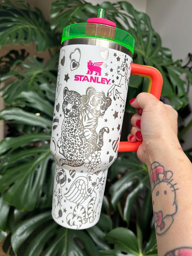 Puede incluir: Un vaso Stanley blanco con una tapa verde ne&oacute;n vibrante, un asa naranja brillante y una pajita a juego de color verde ne&oacute;n. El exterior est&aacute; decorado con ilustraciones de l&iacute;neas negras de un leopardo, un tigre, un unicornio y corazones.