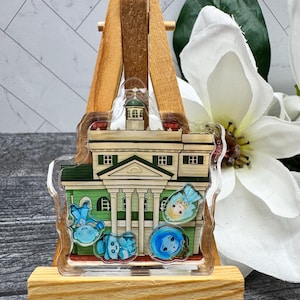 May include: A clear plastic charm featuring a cartoon illustration of the Haunted Mansion from Disneyland. The charm includes four ghosts, one of which is a woman in a blue dress.