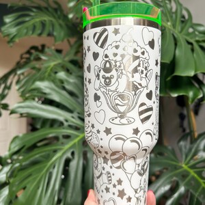 Puede incluir: Un vaso de acero inoxidable blanco con un patr&oacute;n de l&iacute;neas negras de ositos de peluche, helados, globos, corazones y estrellas. El vaso incluye una tapa verde transl&uacute;cida brillante y una pajita verde a juego insertada en la parte superior.