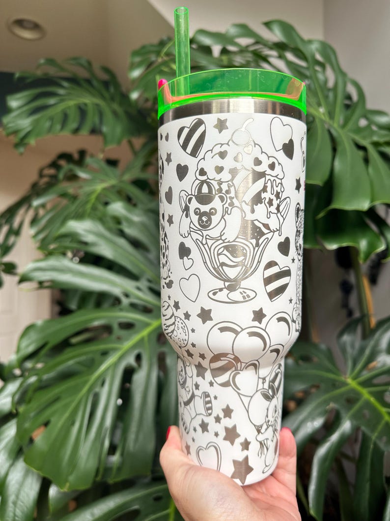 Puede incluir: Un vaso de acero inoxidable blanco con un patr&oacute;n de l&iacute;neas negras de ositos de peluche, helados, globos, corazones y estrellas. El vaso incluye una tapa verde transl&uacute;cida brillante y una pajita verde a juego insertada en la parte superior.