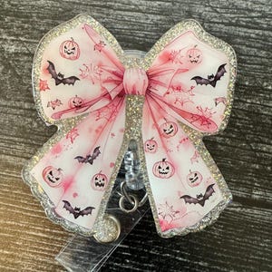 May include: A pink glitter Halloween-themed retractable badge holder with a bow and a design of bats and pumpkins.