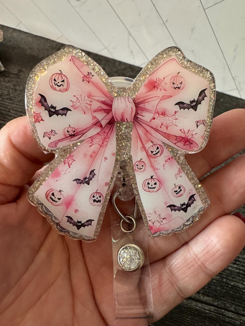 May include: A pink glitter bow with a Halloween theme. The bow features a white background with black bats, orange pumpkins with jack-o-lantern faces, and a spiderweb design. The bow is attached to a clear retractable badge holder with a silver glitter charm.