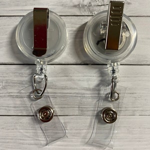 May include: Two clear plastic retractable badge holders with silver clips and a clear plastic strap. The holders are on a white wooden background.