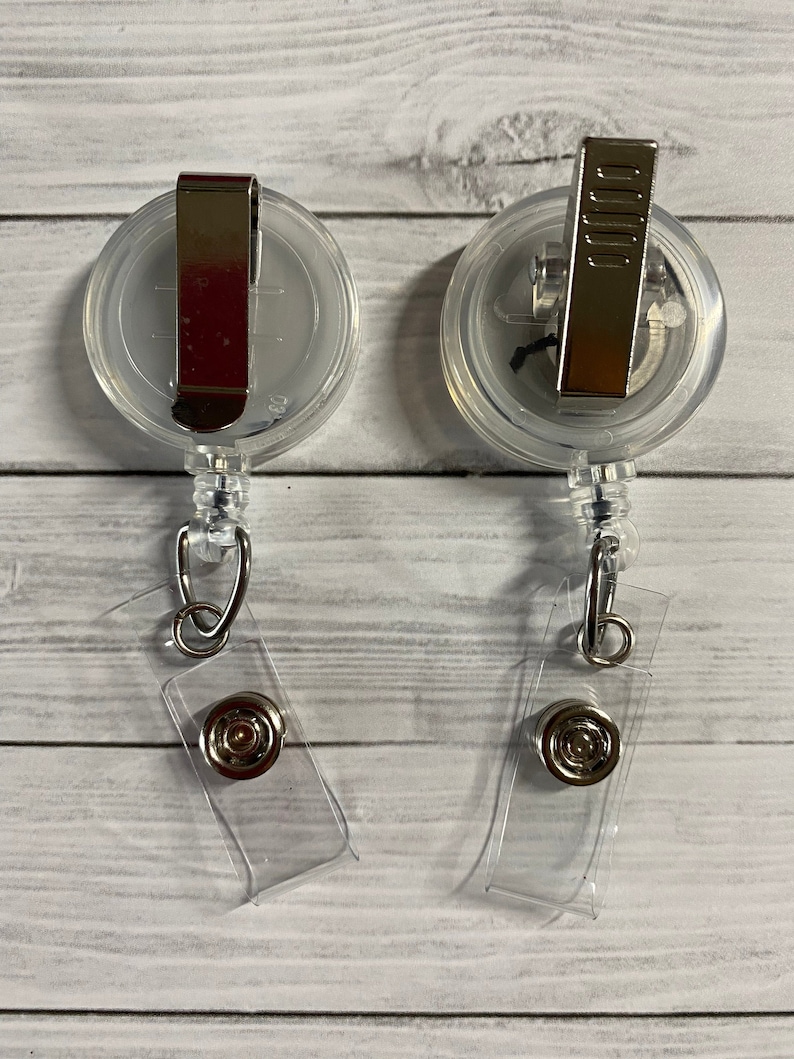 May include: Two clear plastic retractable badge holders with silver clips and a clear plastic strap. The holders are on a white wooden background.