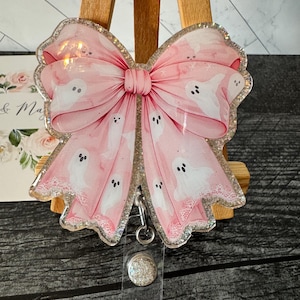 May include: A pink glitter resin badge holder with a white bow and ghost pattern. The badge holder has a clear plastic retractable reel and a sparkly silver button.