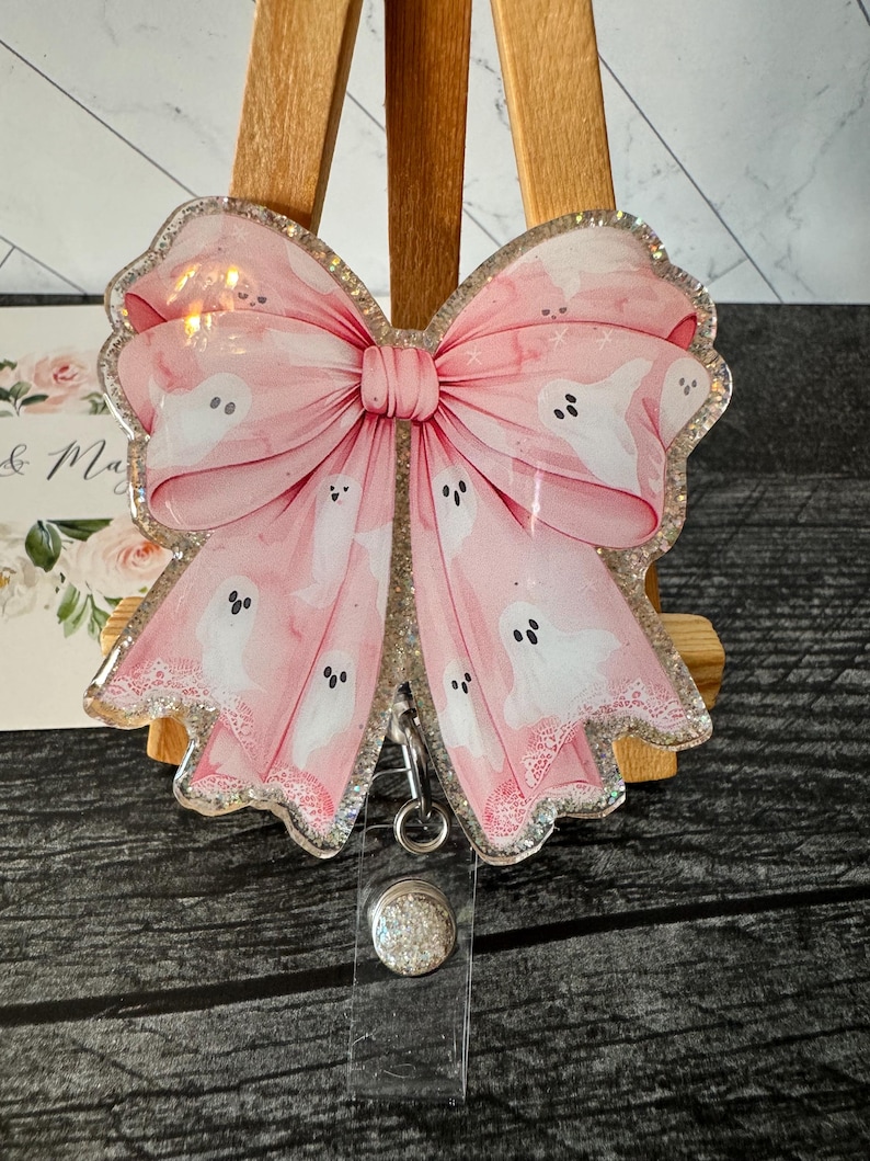 May include: A pink glitter resin badge holder with a white bow and ghost pattern. The badge holder has a clear plastic retractable reel and a sparkly silver button.
