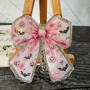 May include: A pink glitter bow keychain with a Halloween theme. The bow is decorated with bats and jack-o'-lanterns.