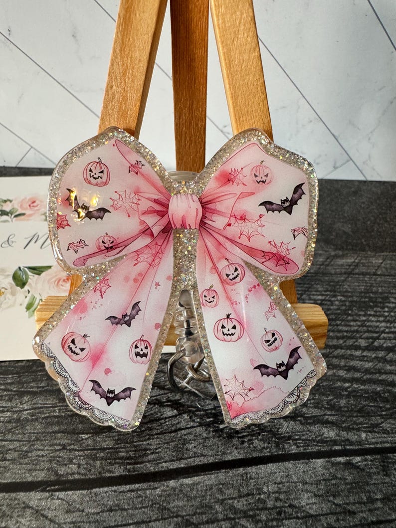 May include: A pink glitter bow keychain with a Halloween theme. The bow is decorated with bats and jack-o'-lanterns.