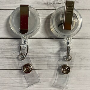 May include: Two clear plastic retractable badge holders with silver clips and clear plastic straps. The holders have a round shape and a clear plastic window for displaying a badge.
