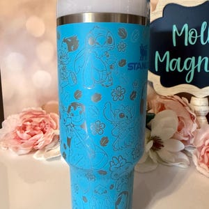 May include: A bright blue Stanley tumbler featuring a repeating laser-etched pattern of Lilo and Stitch characters and tropical flowers. The cup has a white lid with a straw and the Stanley logo is visible on the upper side of the cylindrical body.