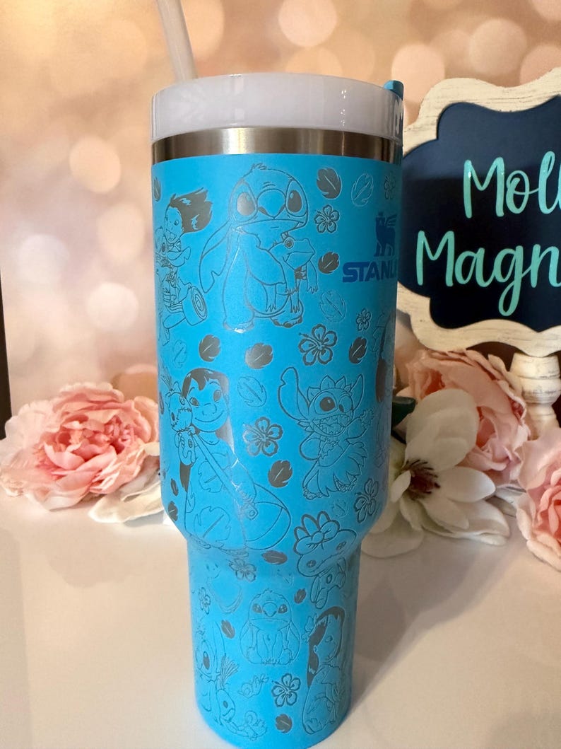 May include: A bright blue Stanley tumbler featuring a repeating laser-etched pattern of Lilo and Stitch characters and tropical flowers. The cup has a white lid with a straw and the Stanley logo is visible on the upper side of the cylindrical body.