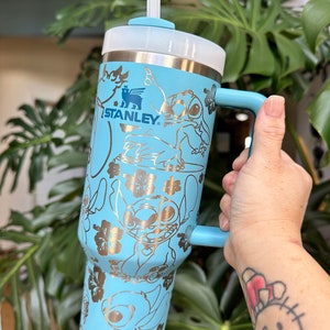 May include: A light blue stainless steel travel mug with a gold metallic design featuring Stitch from Lilo & Stitch. The mug has a handle and a clear lid with a straw. The mug is branded with the Stanley logo.