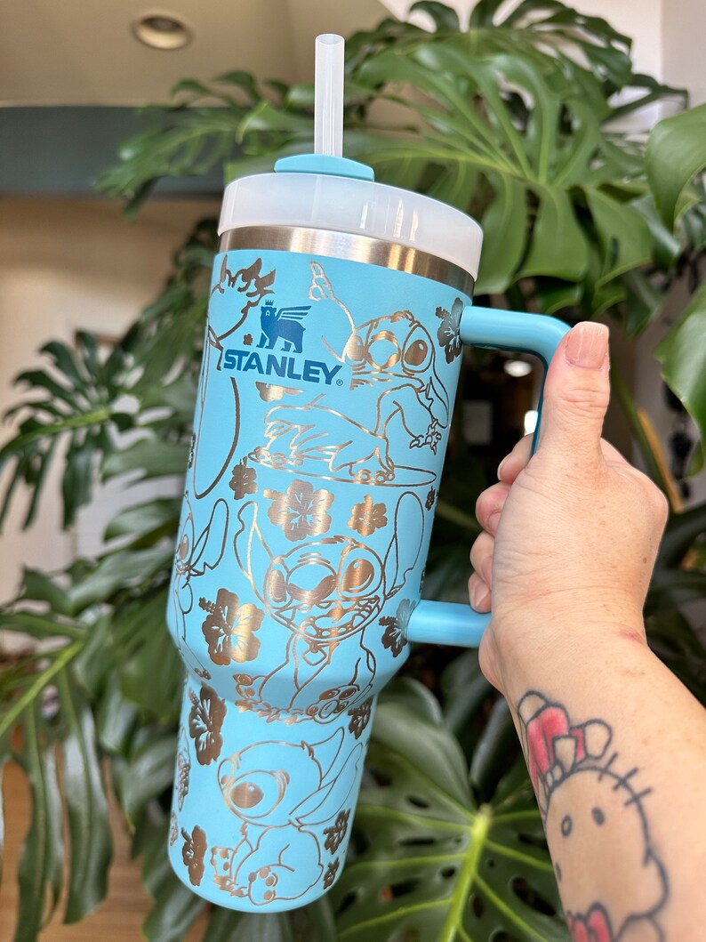 May include: A light blue stainless steel travel mug with a gold metallic design featuring Stitch from Lilo & Stitch. The mug has a handle and a clear lid with a straw. The mug is branded with the Stanley logo.