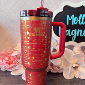 May include: A red Stanley tumbler with a handle, lid, and straw. The tumbler is decorated with gold stars and the Stanley logo. The handle and lid are red. The straw is red and white striped.
