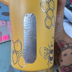 May include: Yellow tumbler with silver bee and honeycomb designs. The tumbler has a silver rectangle on the side. The design includes a bee and honeycomb pattern. The tumbler is held by a person.