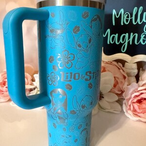 May include: A bright blue insulated tumbler with a large handle and a white lid. The exterior features a repeating etched pattern of Lilo and Stitch characters, tropical flowers, and leaves, with the text Lilo & Stitch printed in the center.