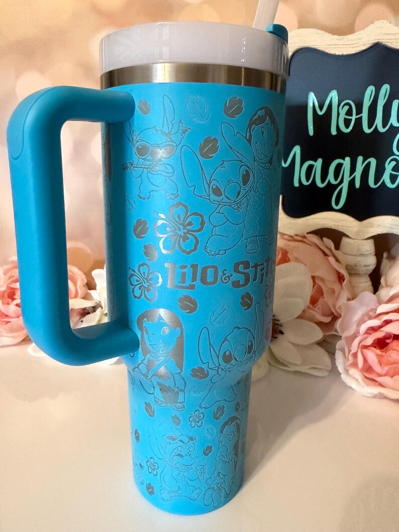May include: A bright blue insulated tumbler with a large handle and a white lid. The exterior features a repeating etched pattern of Lilo and Stitch characters, tropical flowers, and leaves, with the text Lilo & Stitch printed in the center.