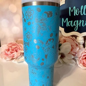 May include: A bright blue stainless steel tumbler featuring a laser-engraved pattern of Lilo and Stitch characters, tropical hibiscus flowers, and leaves. The cup includes a white lid with a blue straw insert and a metallic silver rim.