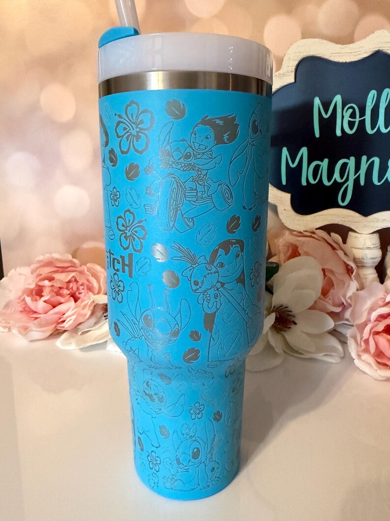May include: A bright blue stainless steel tumbler featuring a laser-engraved pattern of Lilo and Stitch characters, tropical hibiscus flowers, and leaves. The cup includes a white lid with a blue straw insert and a metallic silver rim.