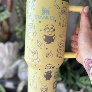 May include: Yellow stainless steel travel mug with a black handle. The mug features a repeating pattern of Minions characters and bananas. The mug has the Stanley logo and the text "STANLEY" on the front.