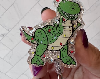 Op de afbeelding: Een groene glitter kerst-dinosaurus met een rode kerstmanmuts en witte kerstverlichting eromheen gewikkeld. De dinosaurus is bevestigd aan een transparante, intrekbare badgehouder van plastic met een groene edelsteen.