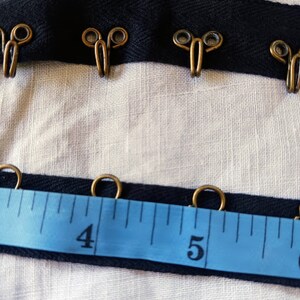 May include: Black ribbon trim with gold metal hooks and loops. The ribbon is measured with a blue tape measure showing the numbers 4 and 5.