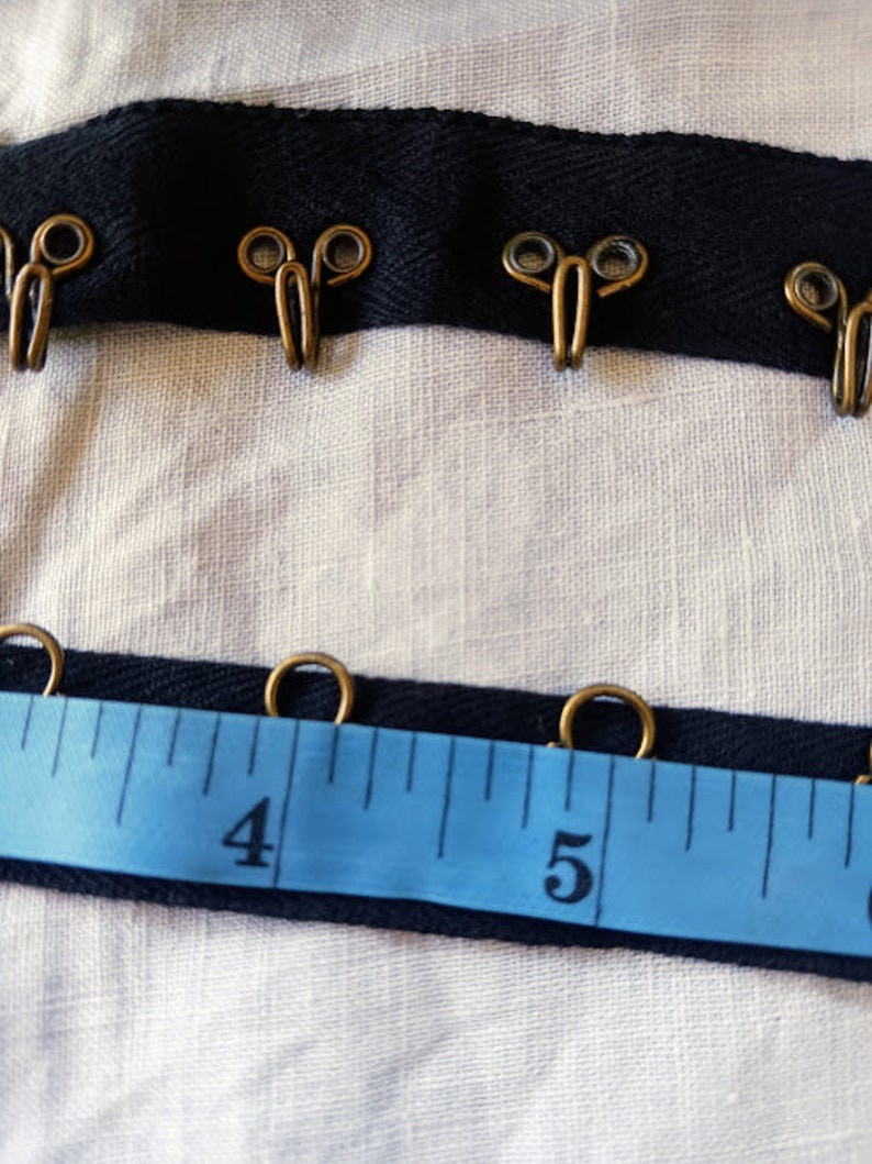 May include: Black ribbon trim with gold metal hooks and loops. The ribbon is measured with a blue tape measure showing the numbers 4 and 5.