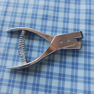 May include: A silver-colored metal hole punch tool featuring a curved handle design and a central coiled spring mechanism. The tool has a hinged construction with two circular rivets visible on the side of the punching head area.