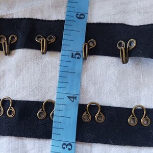 May include: Black ribbon with metal hooks, measured with a blue tape measure. The tape measure shows numbers 3, 4, 5, and 6.