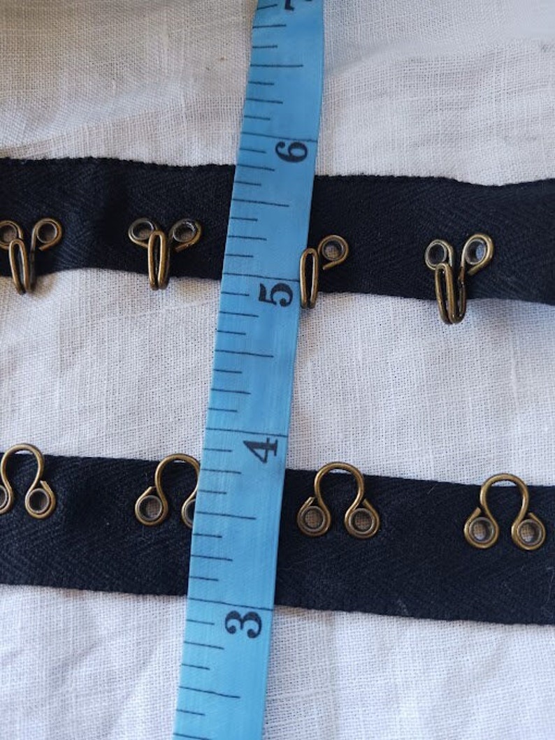May include: Black ribbon with metal hooks, measured with a blue tape measure. The tape measure shows numbers 3, 4, 5, and 6.