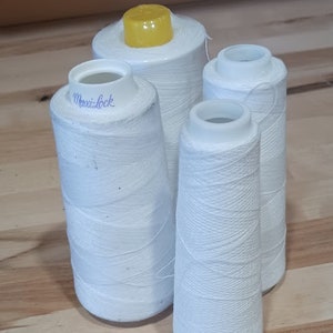 Puede incluir: Cuatro carretes de hilo blanco, uno con la parte superior amarilla y la etiqueta "Maxi-Lock" en el lateral.