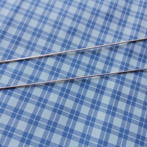 May include: Two silver metal crochet hooks with loops on the ends. The hooks are lying on a blue and white checkered fabric.