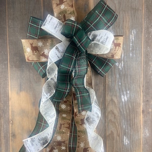 May include: A green plaid ribbon, a white ribbon with a birch bark pattern, and a brown ribbon with a snowflake and deer design are tied together in a bow. The bow is hanging on a wooden surface.