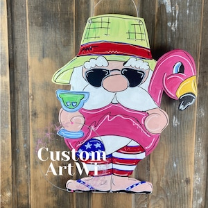May include: A wooden gnome wearing a green and red hat, sunglasses, and a pink flamingo float. The gnome is holding a margarita glass and is wearing red, white, and blue shorts. The gnome is standing on a wooden background.