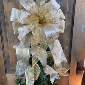 May include: A festive Christmas tree bow featuring layers of white and gold ribbons. The white ribbons are adorned with gold snowflake patterns. The bow is set against a backdrop of a wooden door.