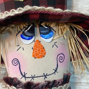 May include: A handmade scarecrow doll with a red and black plaid hat, a burlap face with painted eyes, a nose, and a stitched smile. The doll is wearing a red and black plaid dress with a brown lace trim.