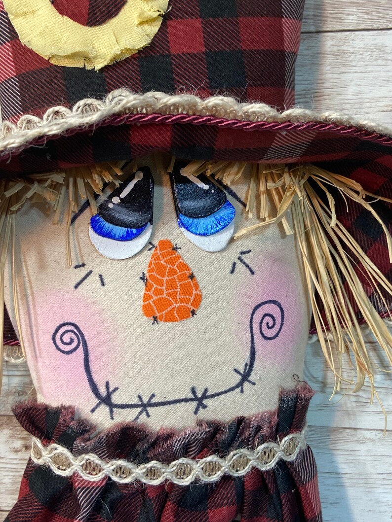 May include: A handmade scarecrow doll with a red and black plaid hat, a burlap face with painted eyes, a nose, and a stitched smile. The doll is wearing a red and black plaid dress with a brown lace trim.