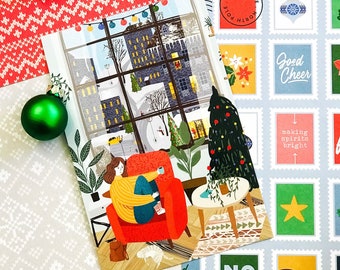 May include: A Christmas card with a winter scene of a person sitting in a red armchair by a window with a Christmas tree in the corner. The card has the text "Good Cheer" and "Noel" on it.