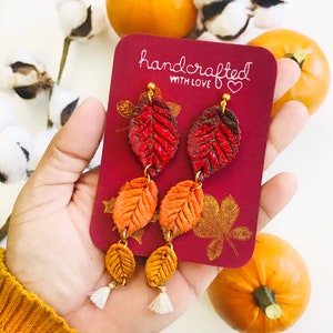 May include: A pair of handmade autumn-themed earrings with three layers of leaves in red, orange, and gold. The earrings are on a red card with the text "handcrafted with love" and "SIZE: LARGE".