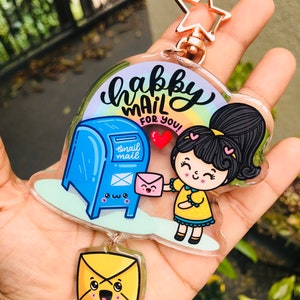 Pu&ograve; includere: Un portachiavi in acrilico trasparente con un design di cassetta postale blu con un viso sorridente e la scritta "snail mail". Una ragazza dei cartoni animati con una coda di cavallo tiene una busta rosa e sorride. Il testo "happy mail for you!" &egrave; scritto in corsivo nero. Il portachiavi ha un ciondolo a forma di stella dorata.