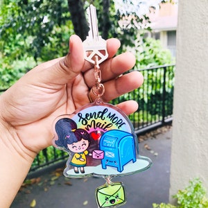 Pu&ograve; includere: Un portachiavi in acrilico colorato con una ragazza dei cartoni animati che tiene una busta rosa e una cassetta delle lettere blu con il testo "Send More Mail" e "Mail Love". Una busta verde con un faccino sorridente &egrave; attaccata alla parte inferiore.