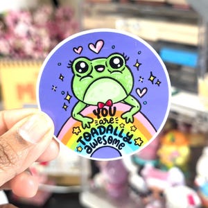May include: A round sticker featuring a cartoon frog with large eyes, a red bow tie, and a rainbow. The text "You are toadally awesome" is written on the sticker. The frog is green, and the background is purple.