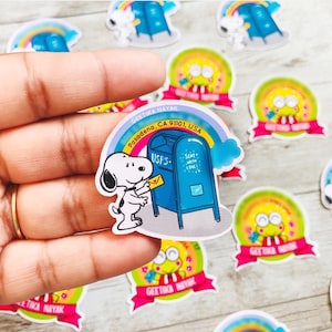 May include: A collection of colorful stickers featuring cartoon characters and designs. One sticker shows Snoopy with a letter at a blue USPS mailbox with the text "Sent with Love!". Other stickers include a rainbow and a character with a pink ribbon.